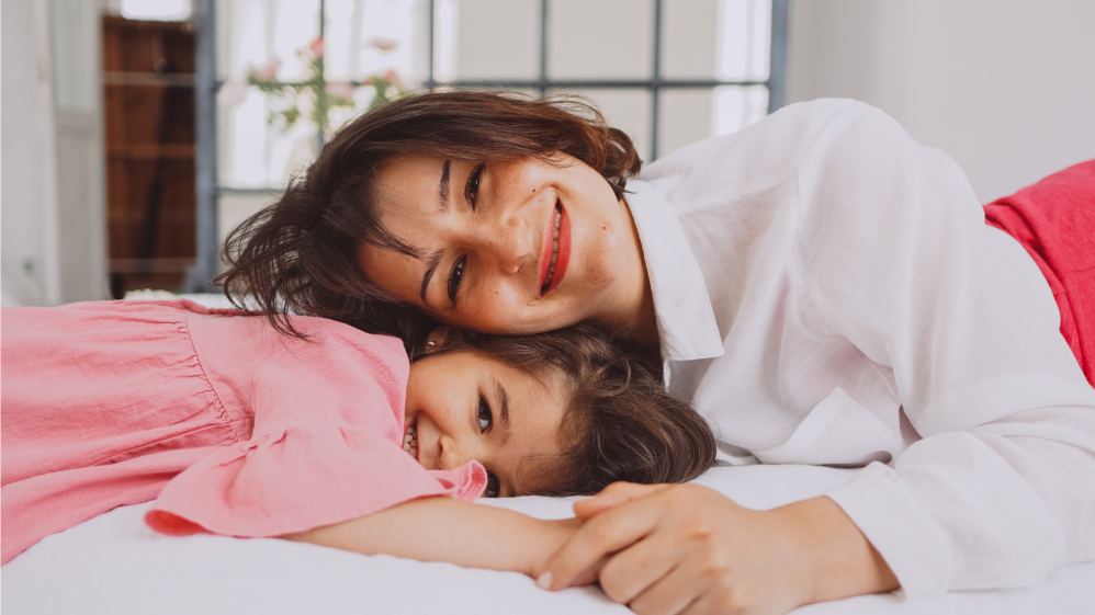 Mãe e filha deitadas na cama, apoiando a cabeça uma na outra, enquanto sorriem para câmera