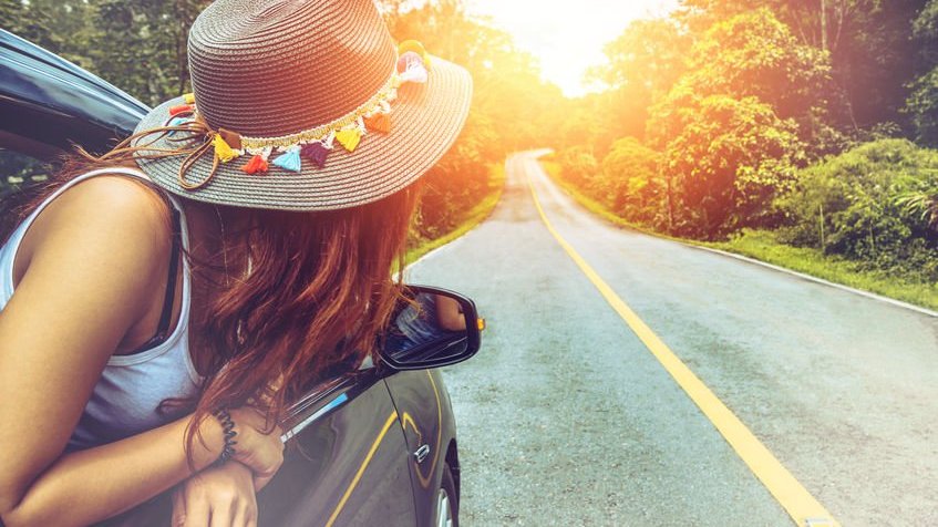 Mulher com meio corpo para fora da janela do carro com chapéu olhando para estrada