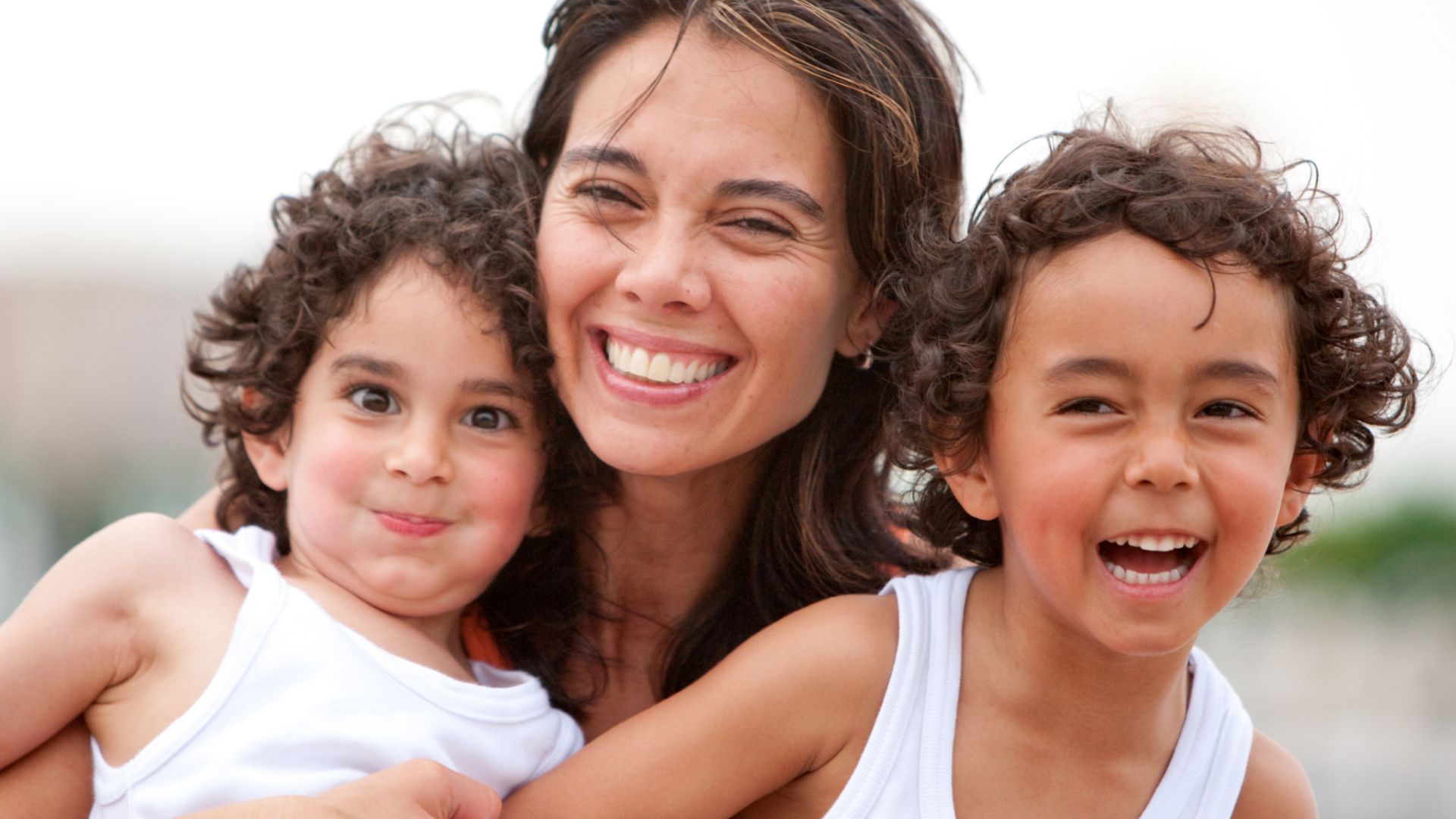 Imagem de uma mãe sorridente posicionada no centro da imagem com os seus dois filhos.