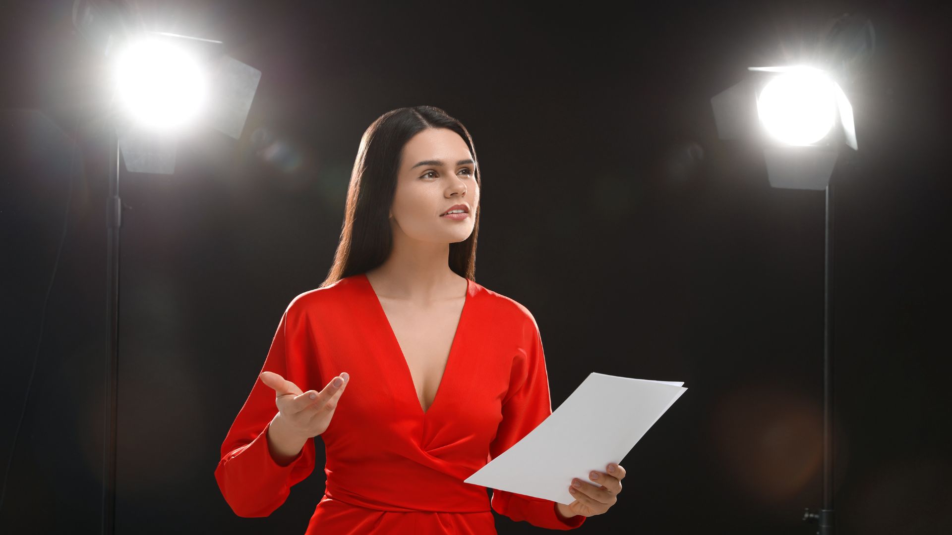 Imagem de um estúdio fotográfico. Em destaque uma atriz usando um vestido vermelho ensaiando o texto para fazer o seu papel.