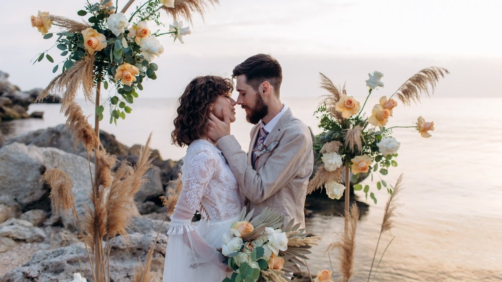 Noivo e noiva em posição de se beijarem, enquanto se casam na praia.