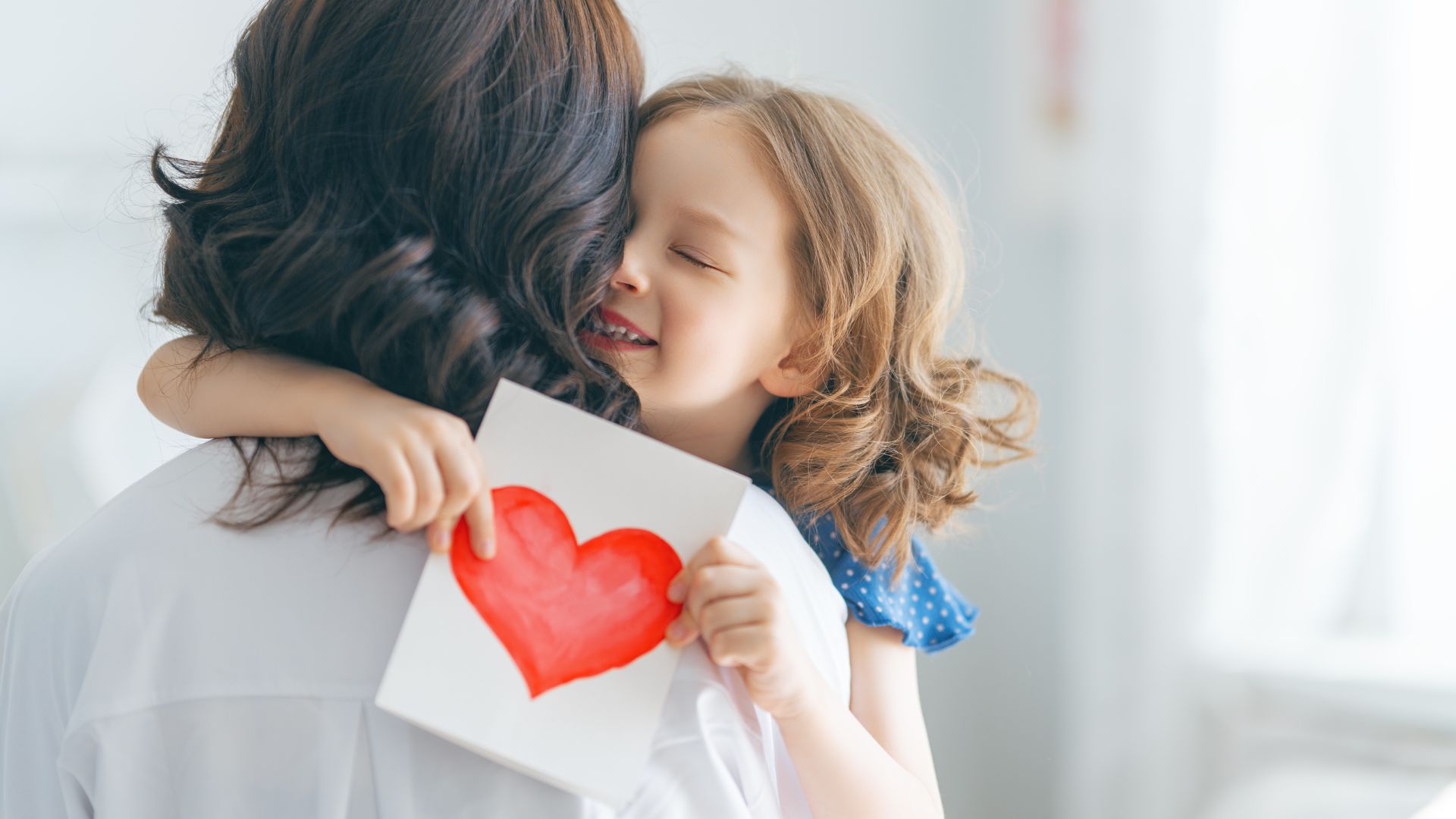 Imagem de uma mãe usando uma camisa branca abrançando a sua filha qe segura uma cartinha com um coração desenhando.