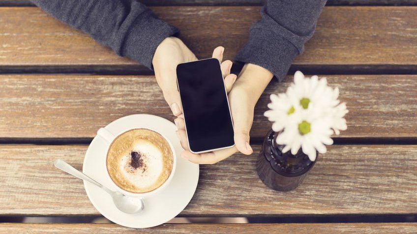 Par de mãos segurando um celular desligado com uma xicara de café e um vaso de flores ao lado