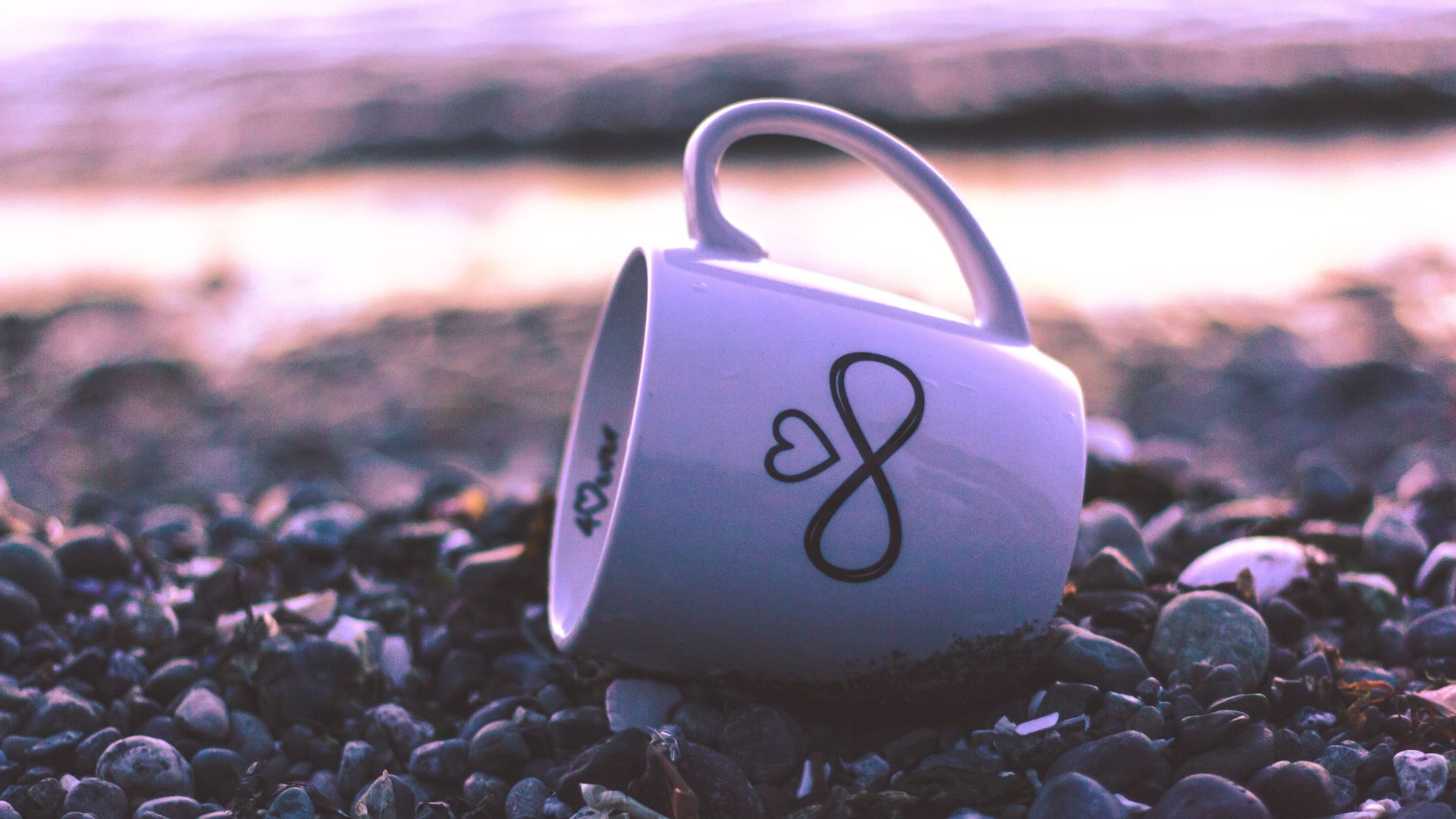 Imagem de um chão de pedras da praia e uma caneca com desenho do infinito e coração nesse chão.