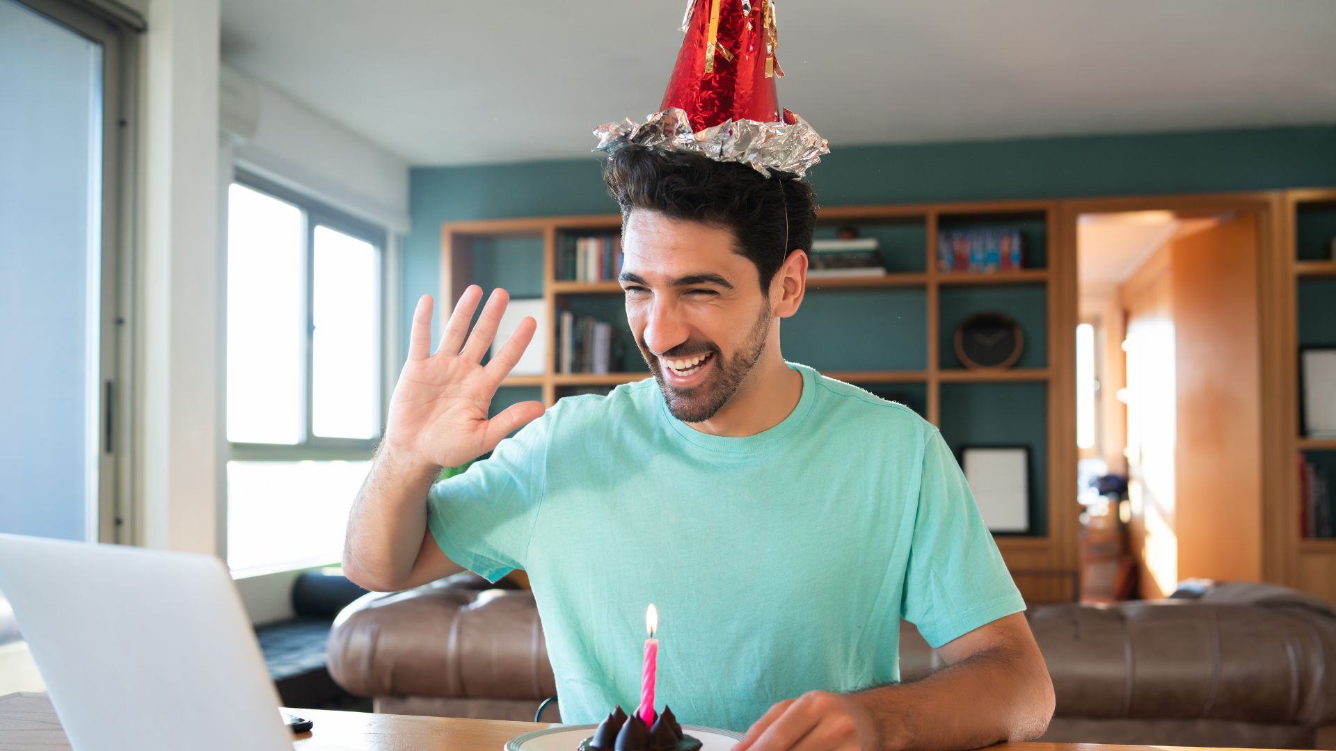 Imagem de um homem saudando e sorrindo para alguém através do notebook com um chapéu de aniversário e um cupcake com uma vela no topo
