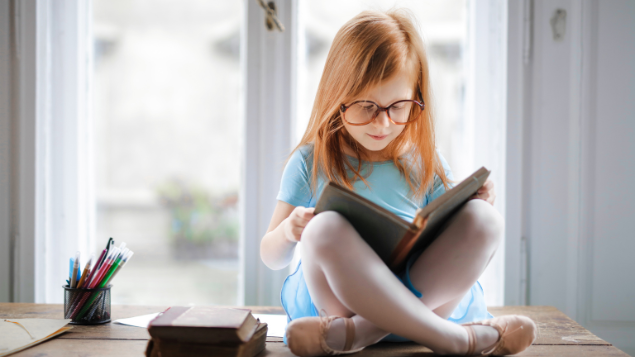 Menina lendo um livro de pernas cruzadas perto de uma janela