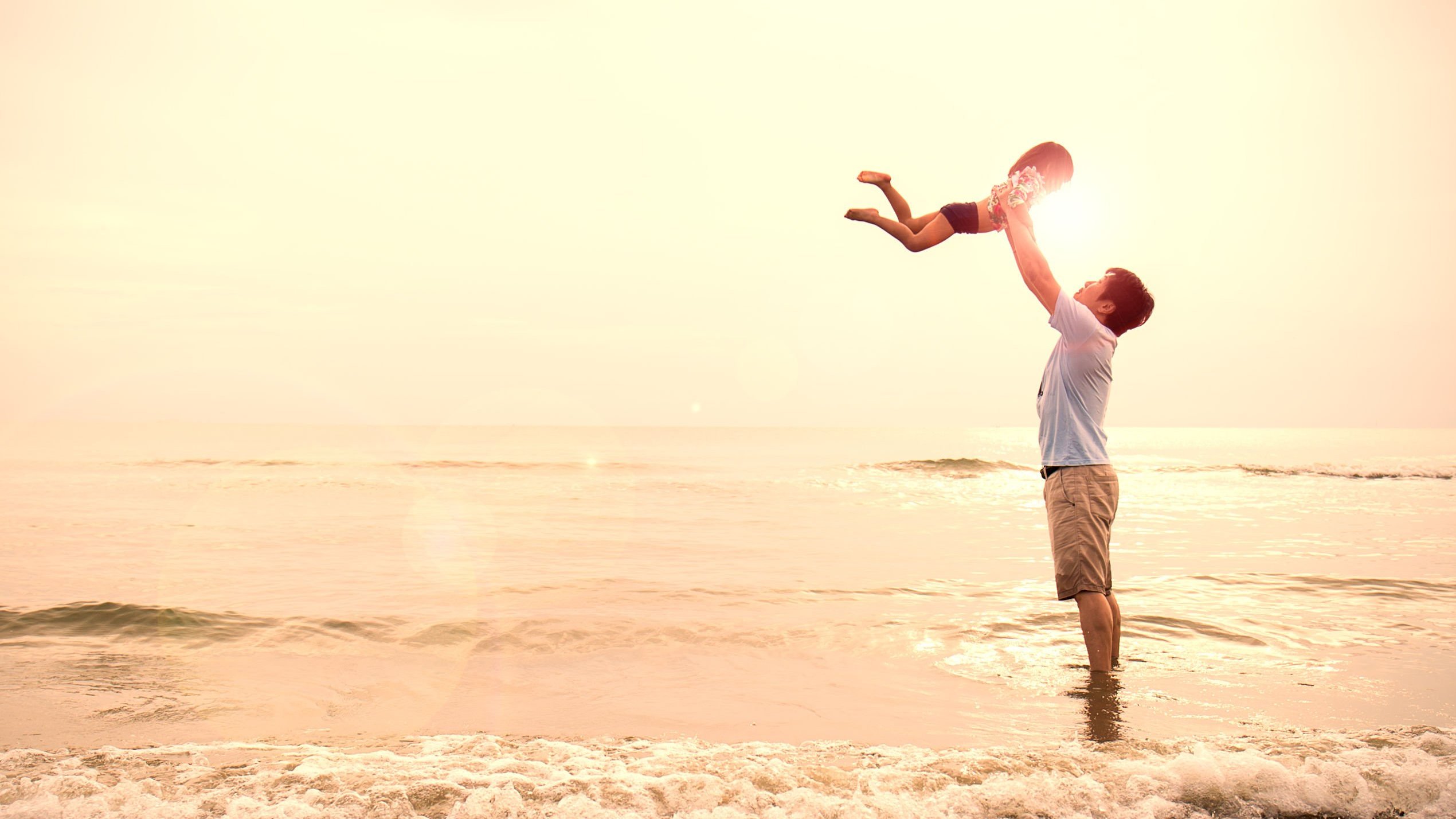 Homem segurando menina pequena para o alto, com os pés no mar em uma praia, com o sol ao fundo.