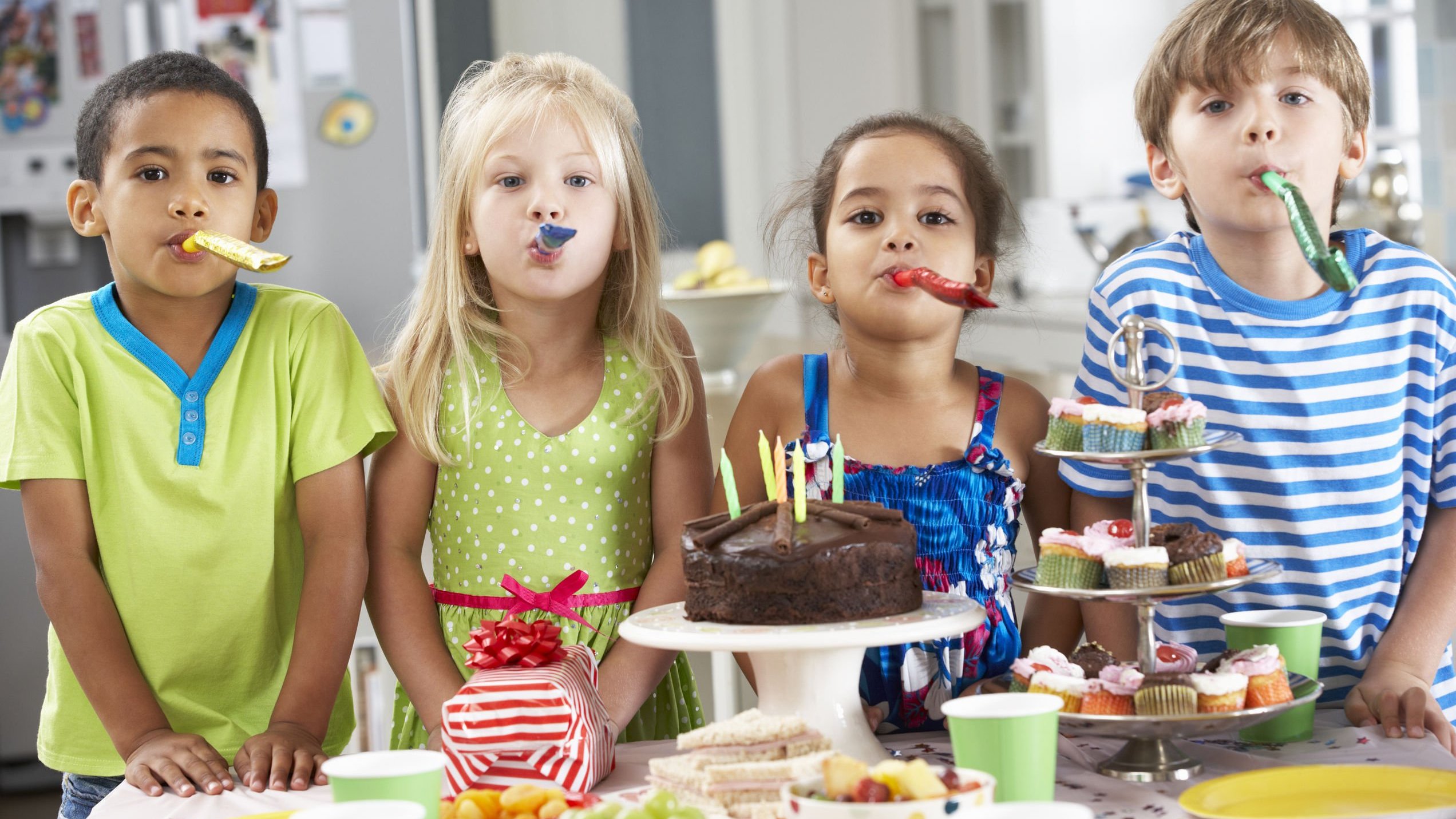 Quatro crianças lado a lado atrás de uma mesa de festa de aniversário, assoprando línguas-de-sogra.