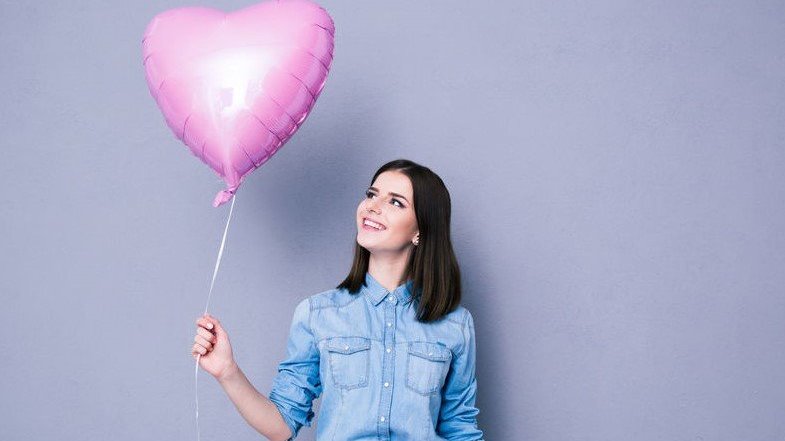 Mulher jovem segurando balão rosa em formato de coração e olhando para ele.