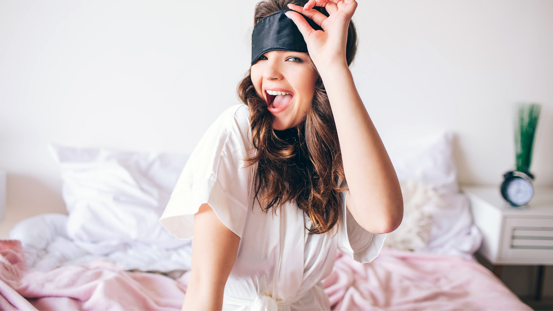 Mensagens Com Amor / Canva / Anton Estrada Imagem de uma moça que acabou de acordar sentada na cama, sorrindo e tirando o tapa olho para dormir.