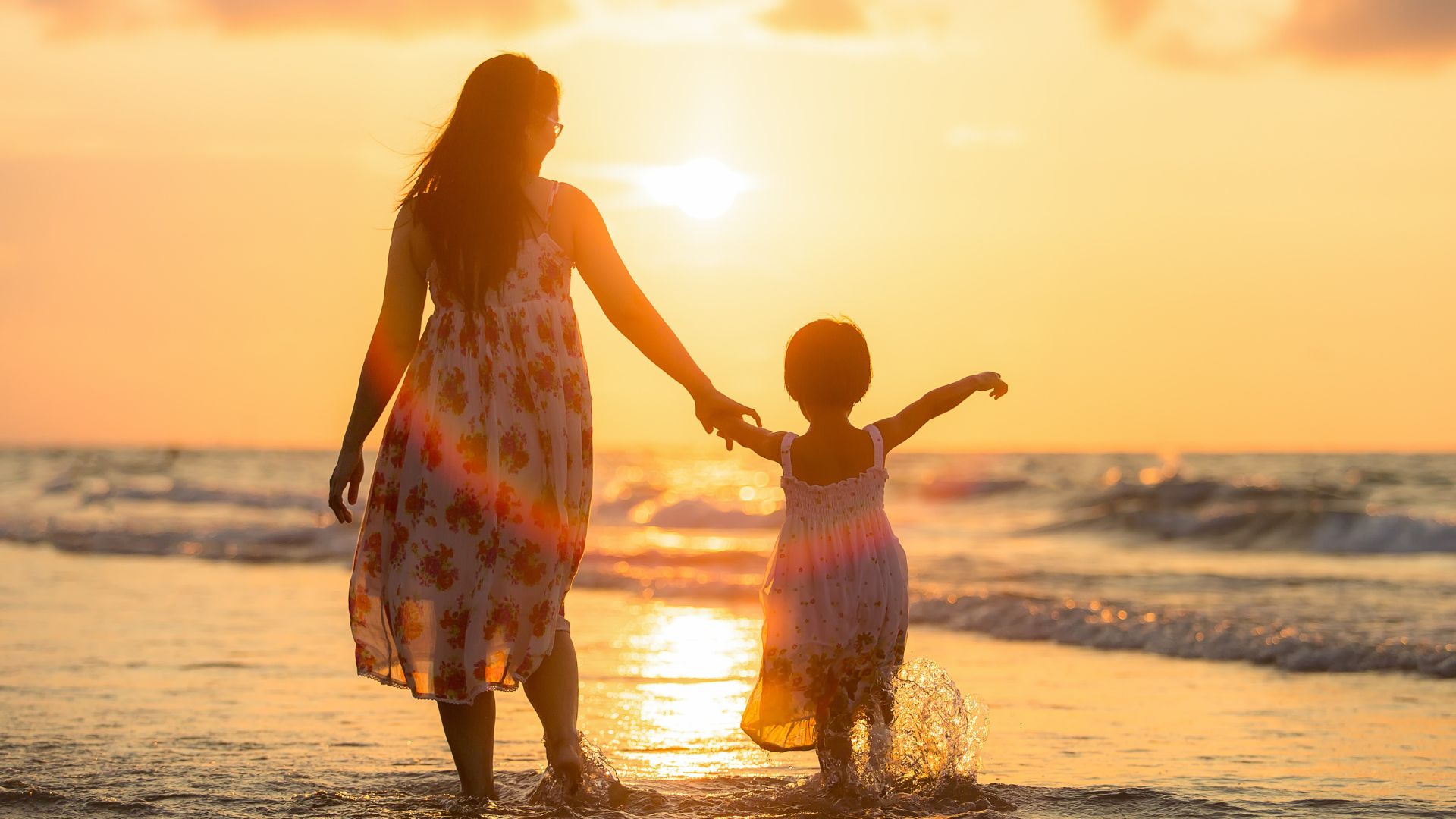 Imagem do mar e ao fundo um lindo pôr do Sol. Em destaque, mãe e filha de mãos dadas, caminhando na areia da praia. Ambas usam vestido branco com flores.