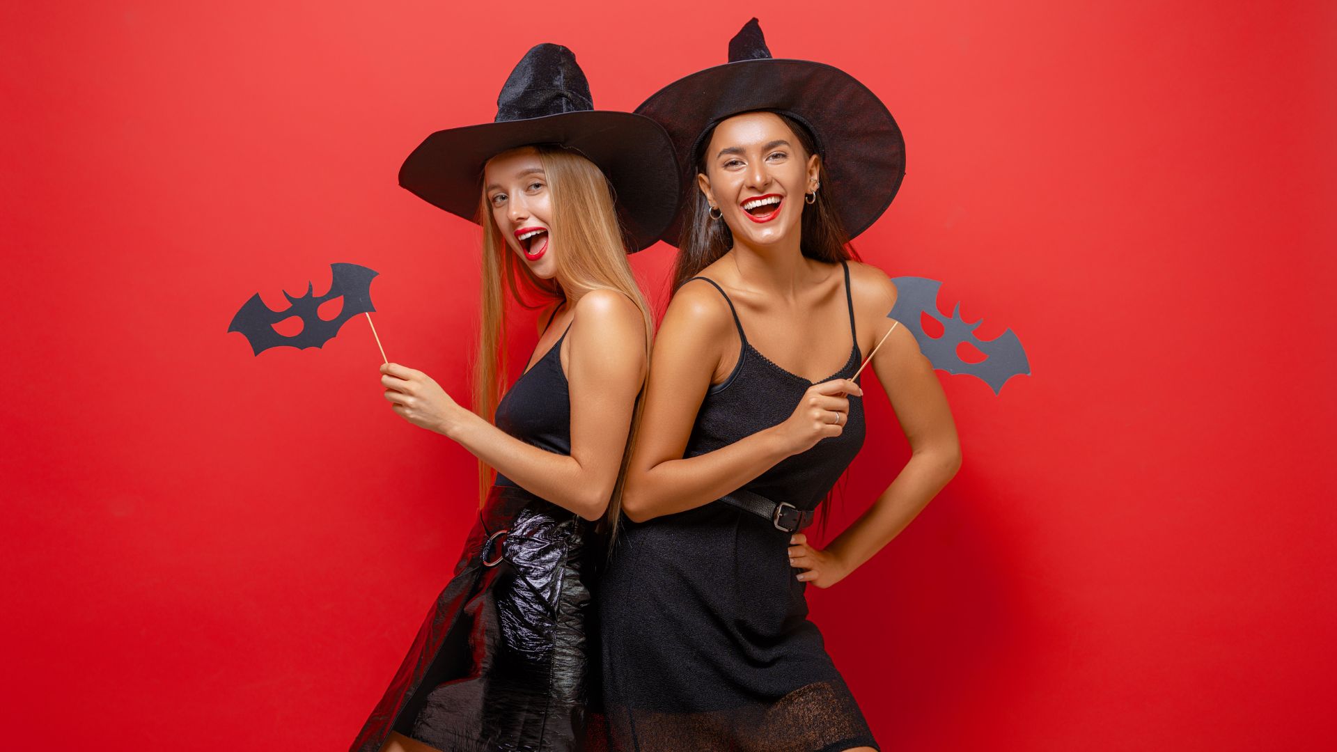 Imagem de fundo vermelho com duas mulheres (uma loura e outra morena) fantasiadas de bruxas. Elsas usam vestidos e chapéus pretos e seguram uma máscara de morcego.