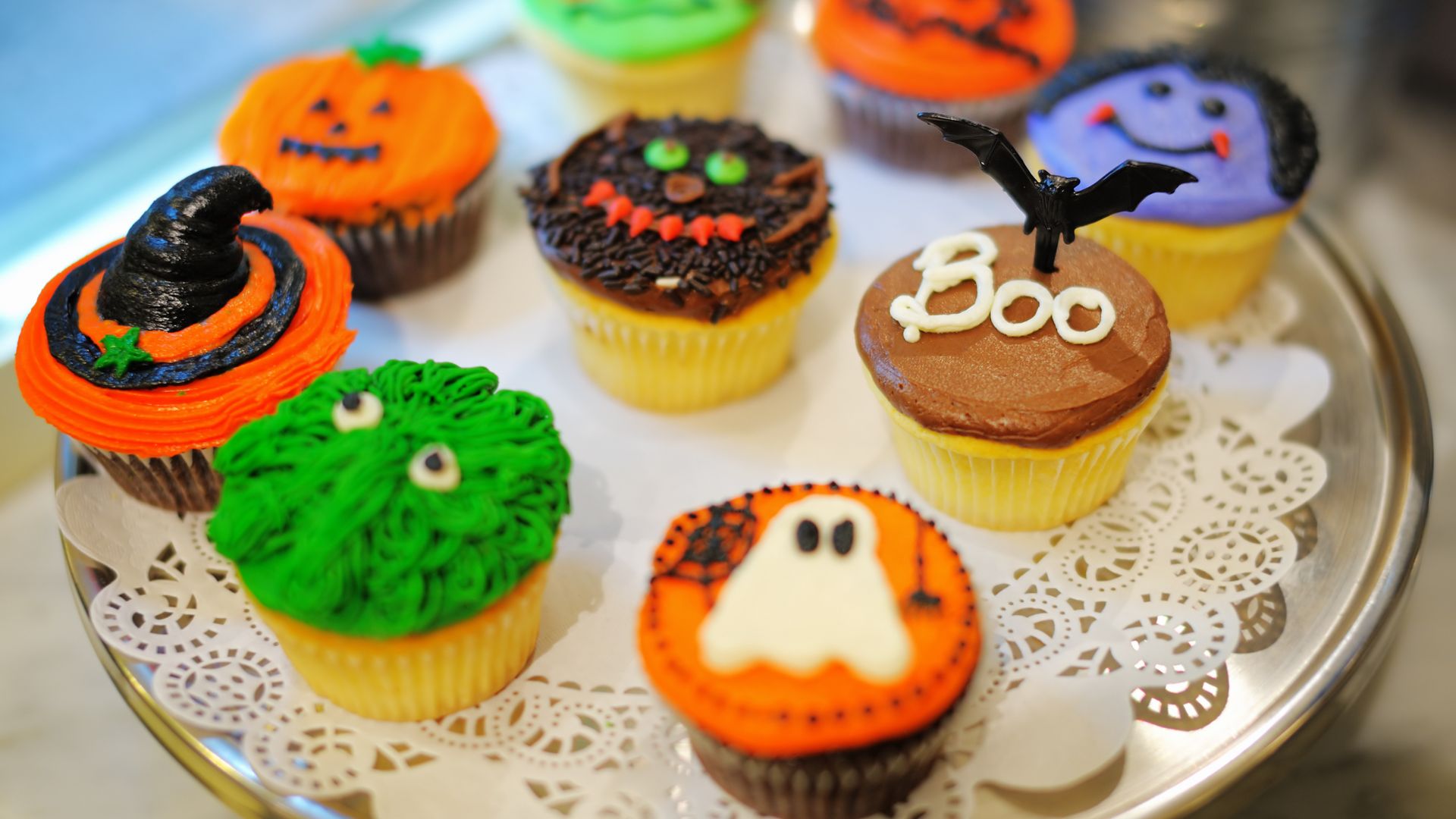 Imagem de vários Cupcakes decorados com imagens de Halloween. Eles estão dispostos em numa bandeja de prata sobre uma toalha decorativa na cor branca.