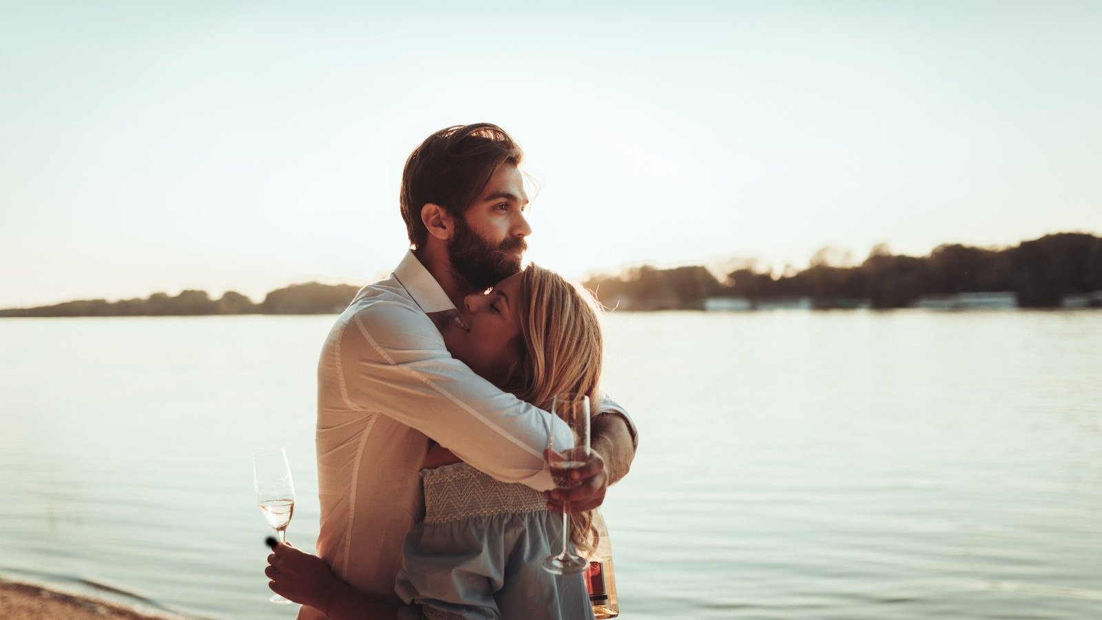 Homem e mulher brancos abraçados próximo ao rio, com taças na mão.