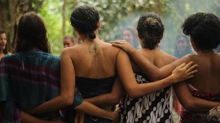 Grupo de mulheres de costas para a câmera, na natureza. Elas estão abraçadas e formam um círculo.