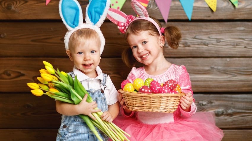 Menina e menino pequenos com orelhas de coelho. O menino segura um buquê de flores, enquanto a menina segura uma pequena cesta cheia de ovos coloridos.