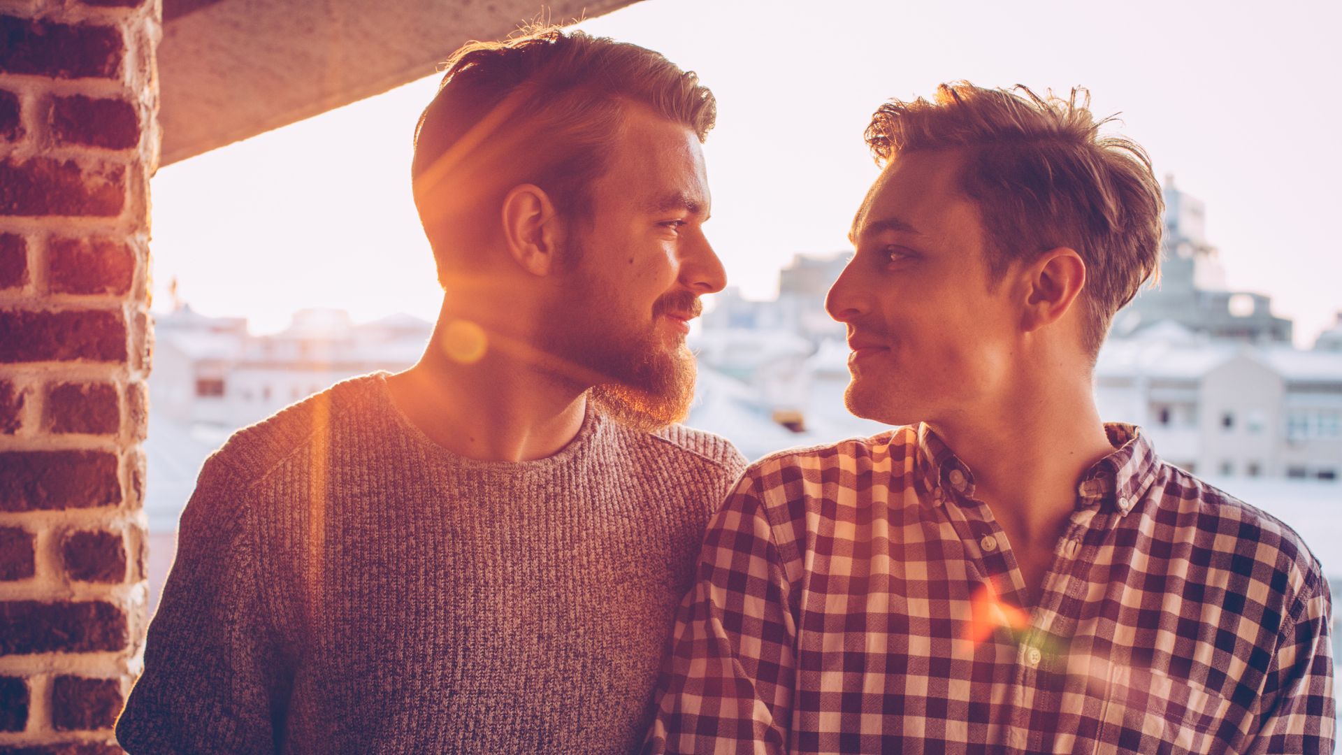 Imagem de um casal gay se olhando e sorrindo e o sol refletido atrás deles
