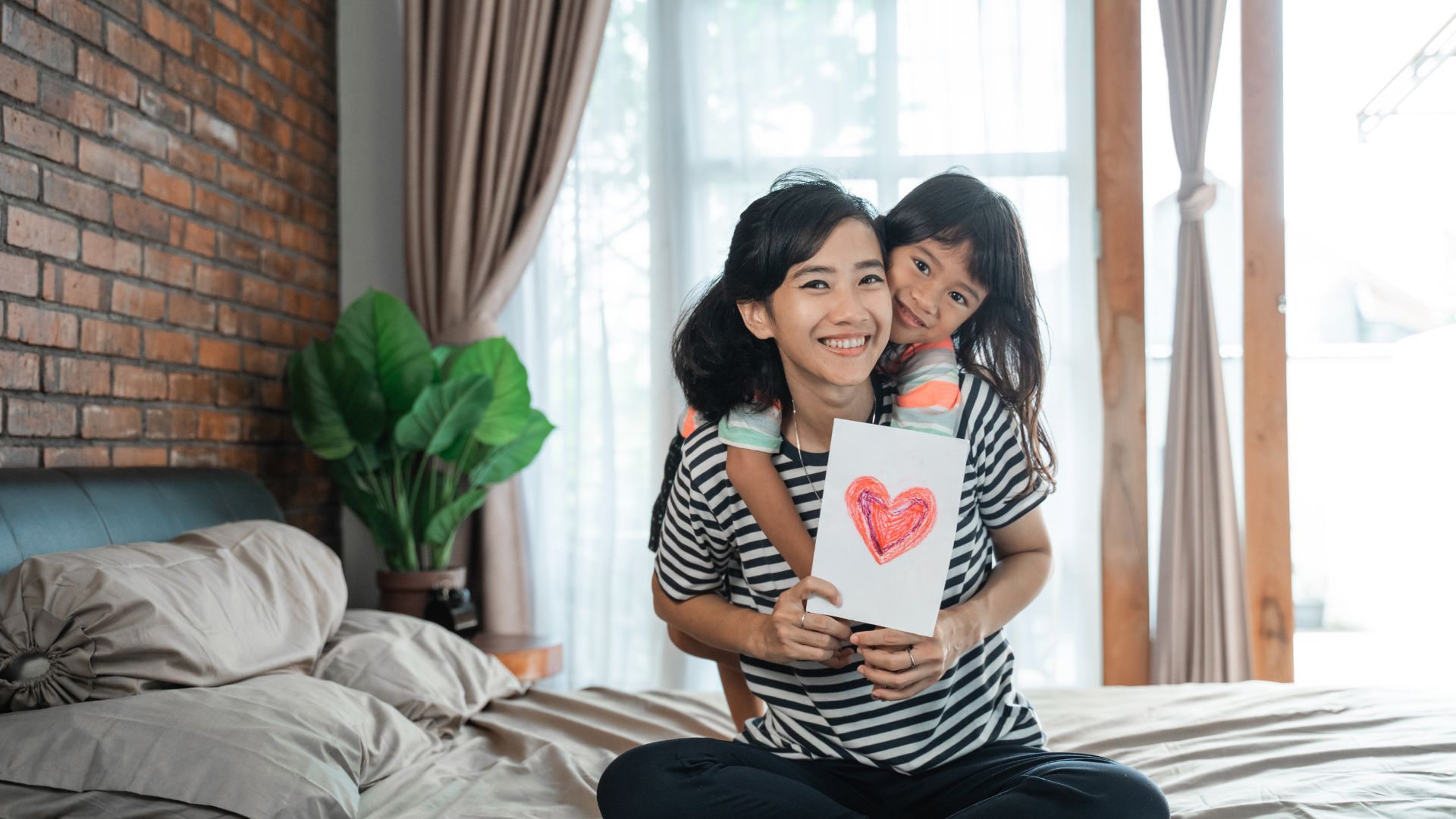 Imagem de uma mãe sentada em uma cama, segurando uma carta com desenho de coração. Ela usa camiseta listrada nas cores branco e preto e está sendo abraçada por sua filha.