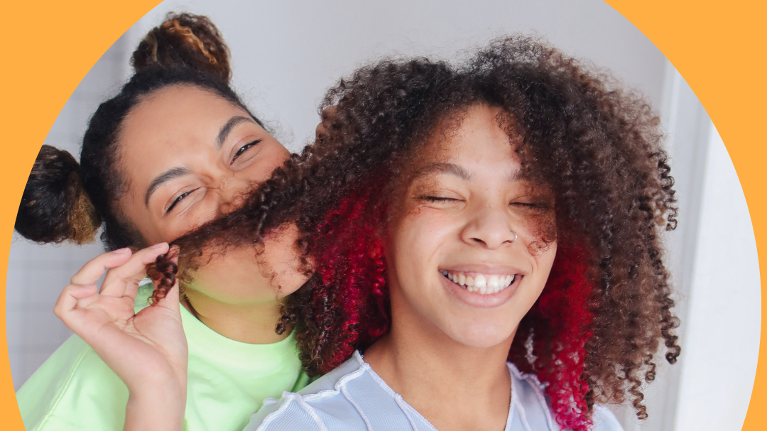 Foto da mãe segurando e cheirando os cabelos cacheados da filha adolescente, que está sorrindo.