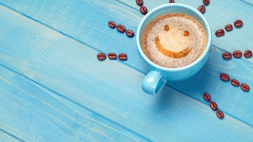 Xicara de café com um sorriso na bebida e com grãos de café formando raios de sol ao seu redor em uma mesa azul