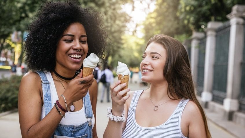 Garotas felizes tomando casquinha de sorvete com sol ao fundo refletindo em calçada