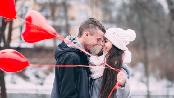 Um homem e uma mulher se abraçando enquanto a mulher segura balões vermelhos em formato de coração
