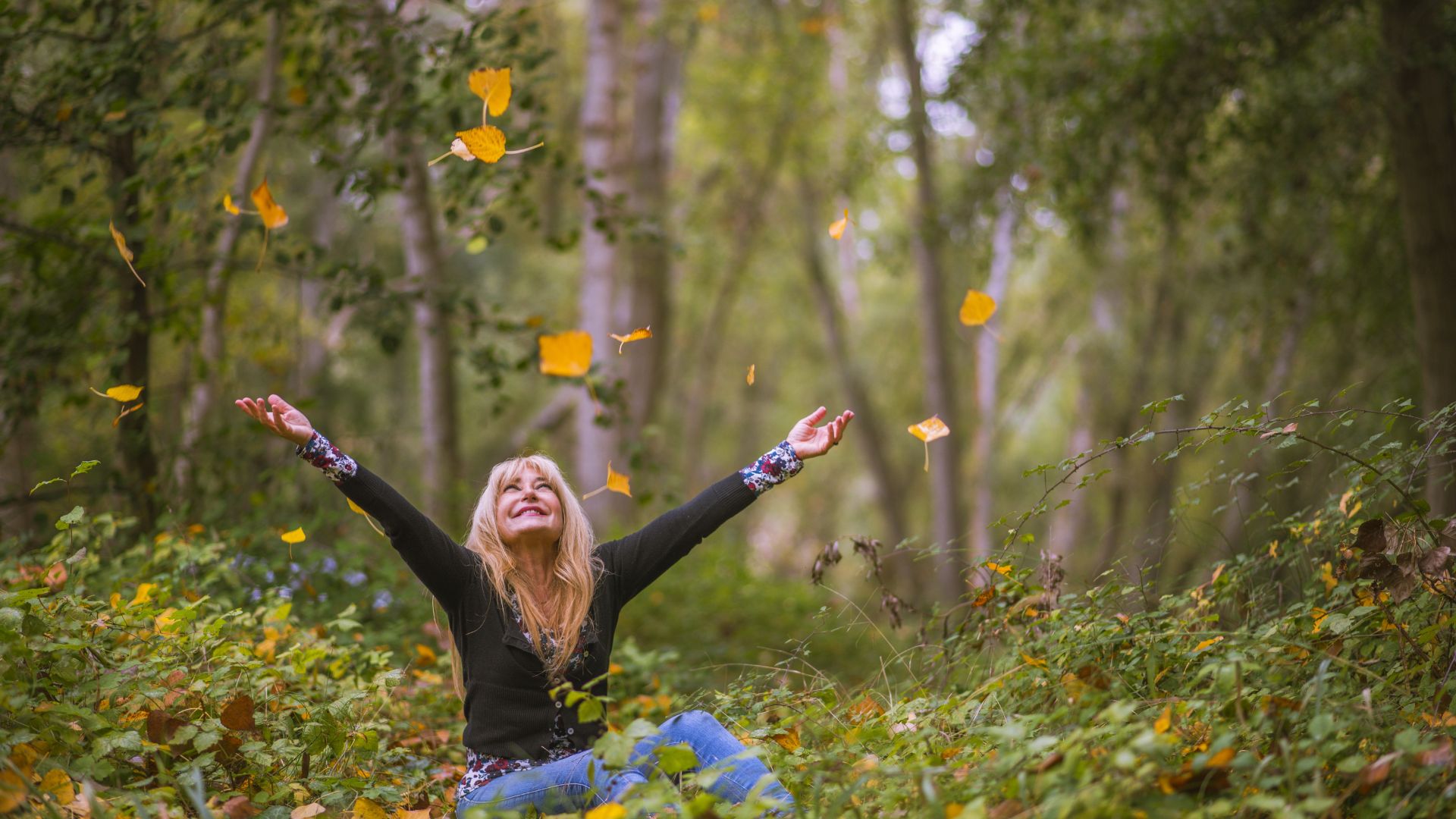 Imagem de uma floresta e ao contro uma mulher loura, de cabelos longos, sentada no chão, sorrindo e feliz.