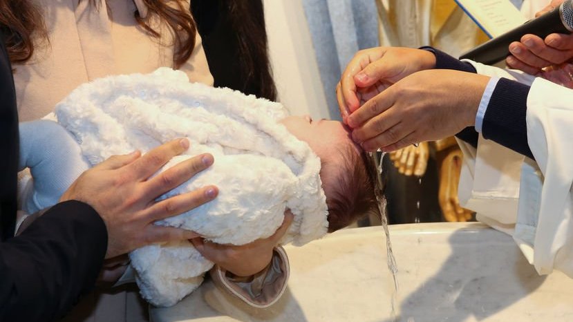 Bebê no colo da mãe, recebe água benta das mãos de um padre em sua cabeça.