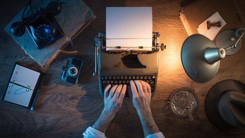 Mãos digitando em máquina de escrever com luminária iluminando a mesa vista de cima