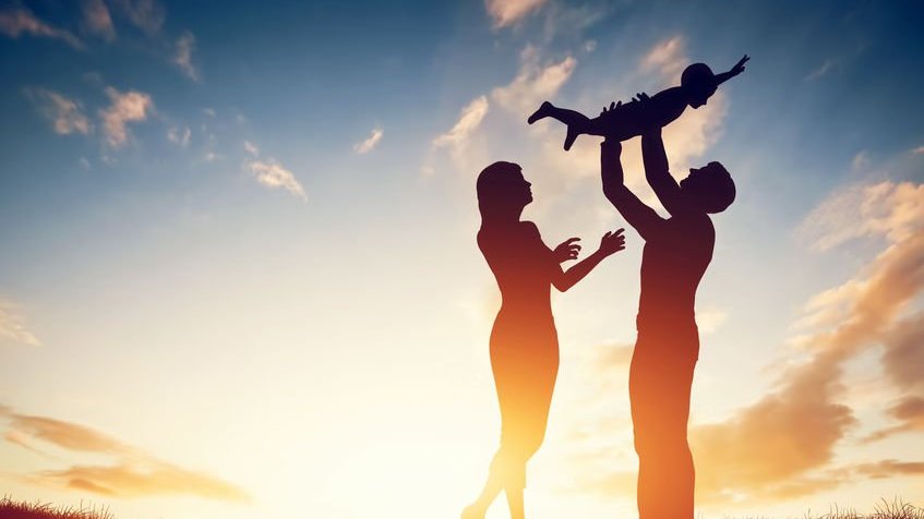 Homem e mulher em ambiente externo seguram uma criança no alto.