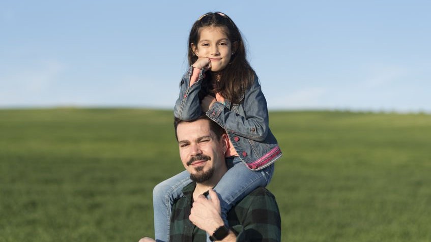 Homem carregando menina em seus ombros, e a pequena apoia com o cotovelo na cabeça do adulto.