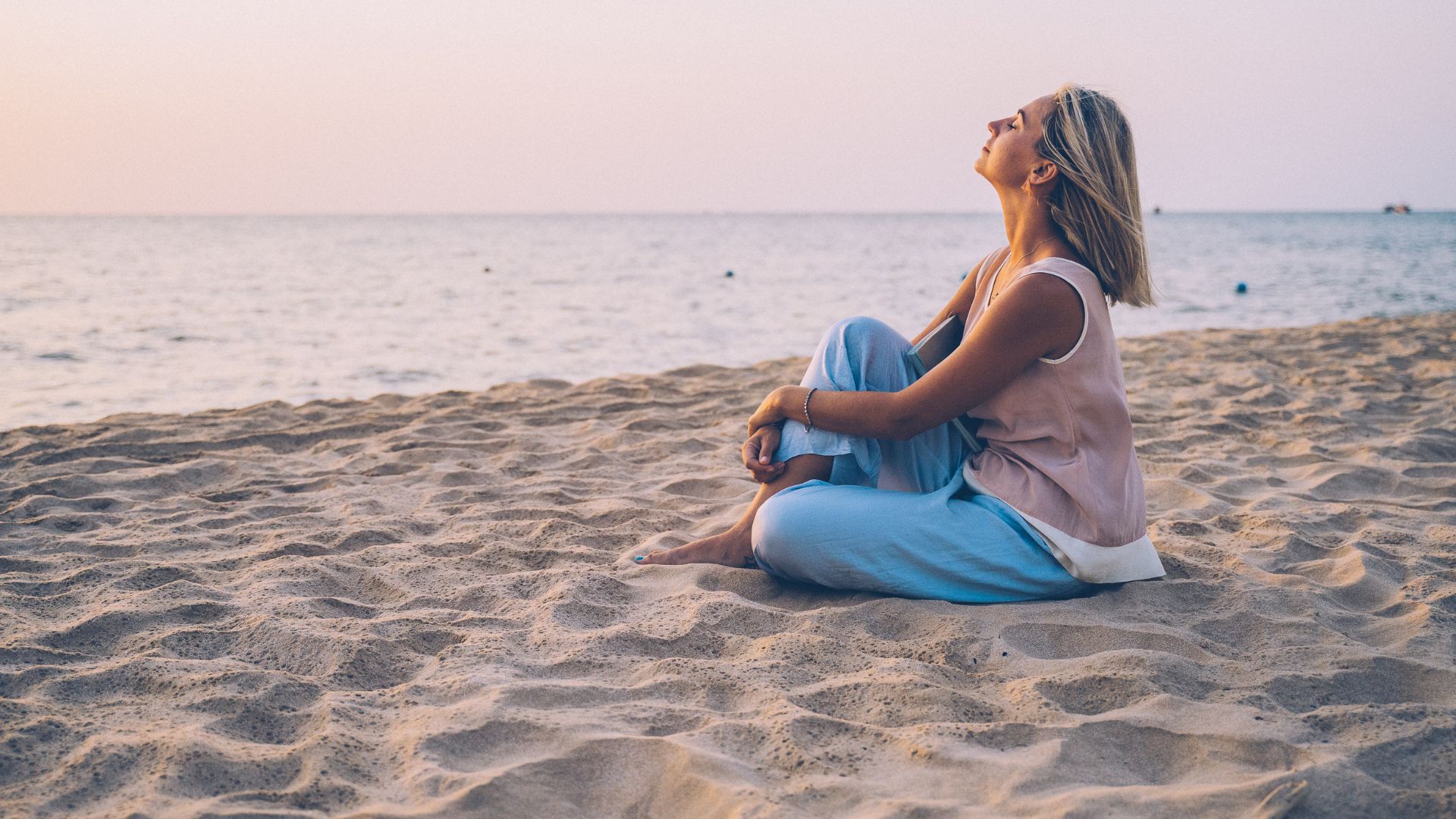 Imagem de uma praia com uma mulher de cabelos curtos sentada sobre a areia. Ela está reflexiva e pensativa por tudo que já superou na vida.
