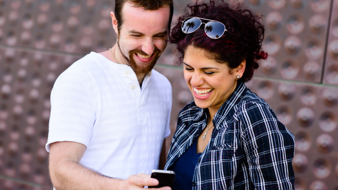 Homem e mulher rindo enquanto estão olhando para o celular.
