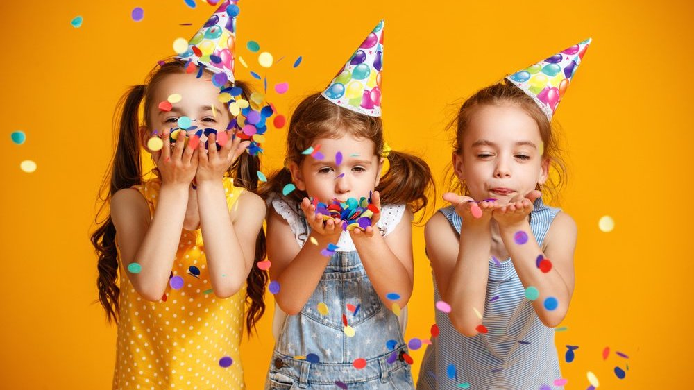 Três meninas usando chapéus de festa assoprando confetes no ar
