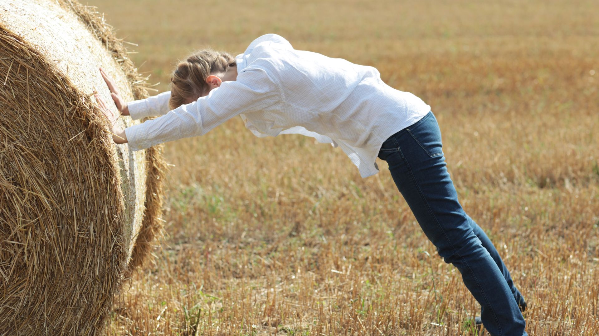 Imagem de uma mulher usando uma camisa branca e calça jeans. Ela está em um campo, empurrando um grande maço de feno.