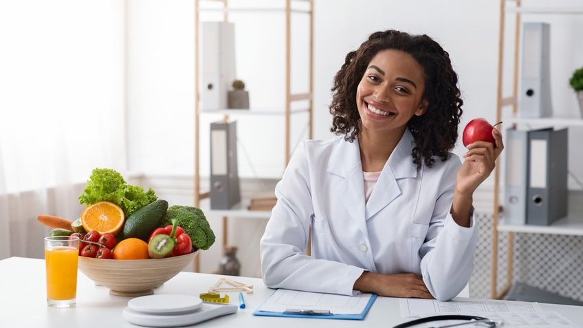 Mulher negra de jaleco segurando maçã num consultório.