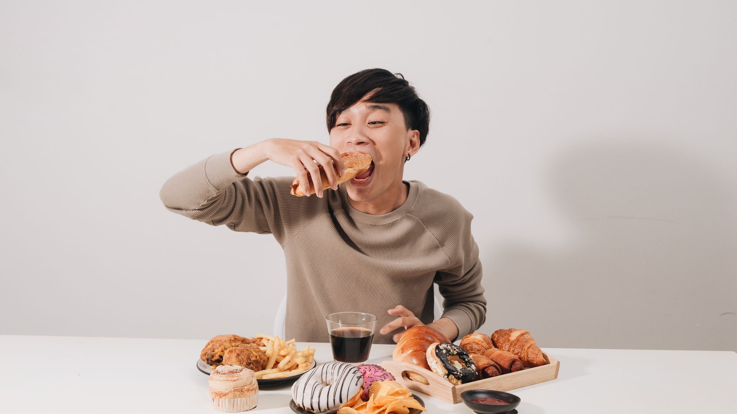 Homem asiático sentado em mesa comendo donuts, salgadinhos, batatas fritas e comidas de fast food.