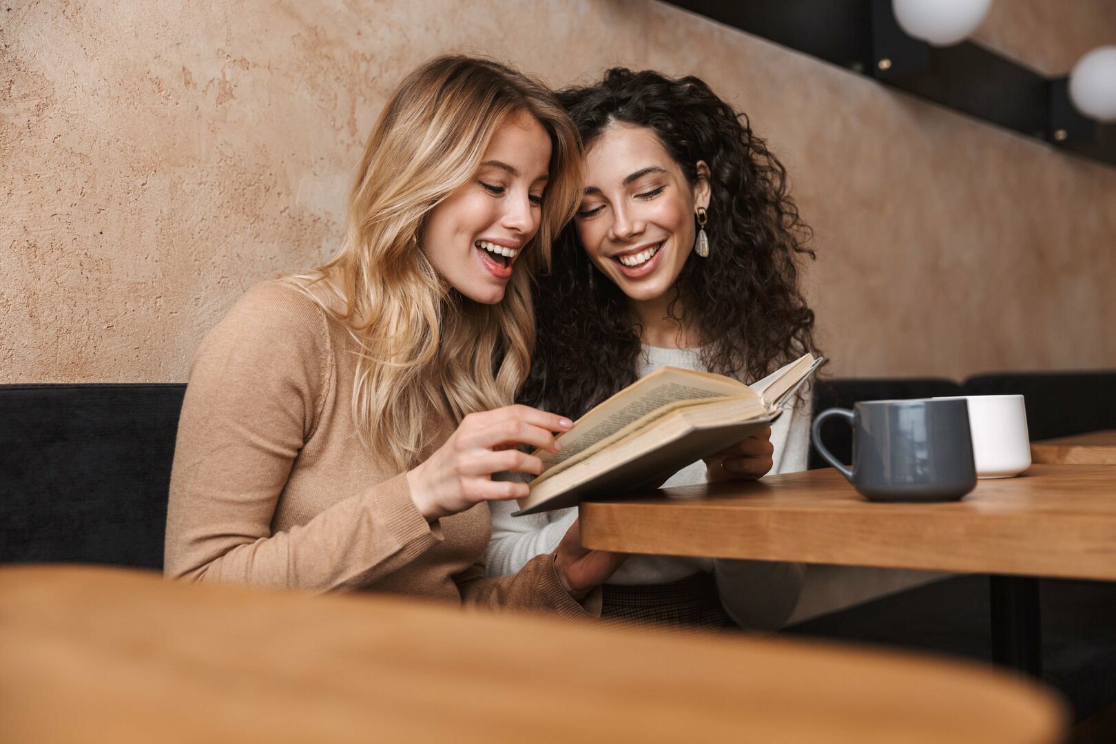 Amigas lendo um livro e sorrindo em frente a uma mesa