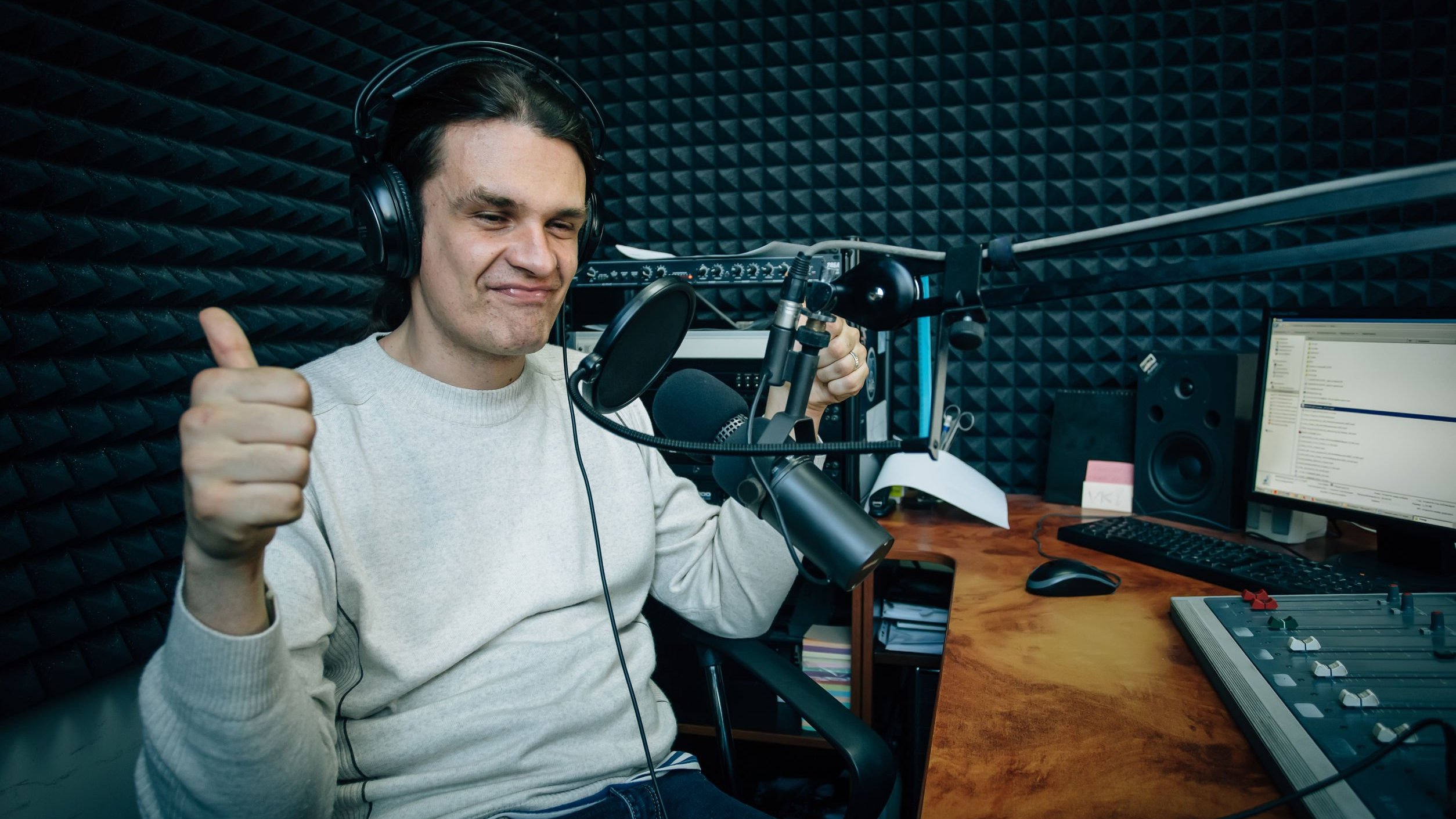 Homem branco com polegar levantado, usando fones de ouvidos e elementos eletrônicos.