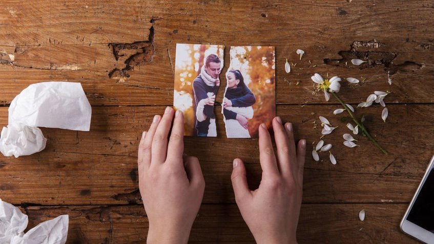 Mãos de uma mulher segurando uma foto rasgada de um casal, ao lado de flores murchas e lenços de papel amassados