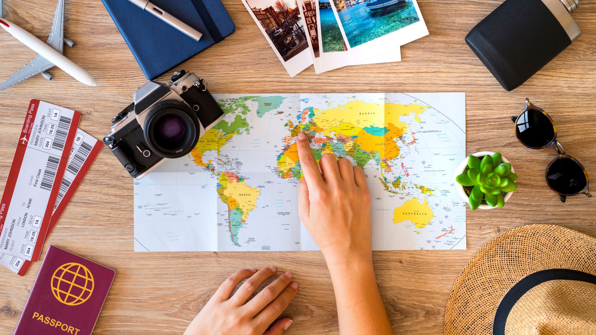 Imagem de uma mesa de madeira. Sobre ela, um amão feminina apontando lugares no mapa mundo. Passaporte, passagens, chapeú, miniatura de avião, fotos, óculos de sol e máquina fotográfica.