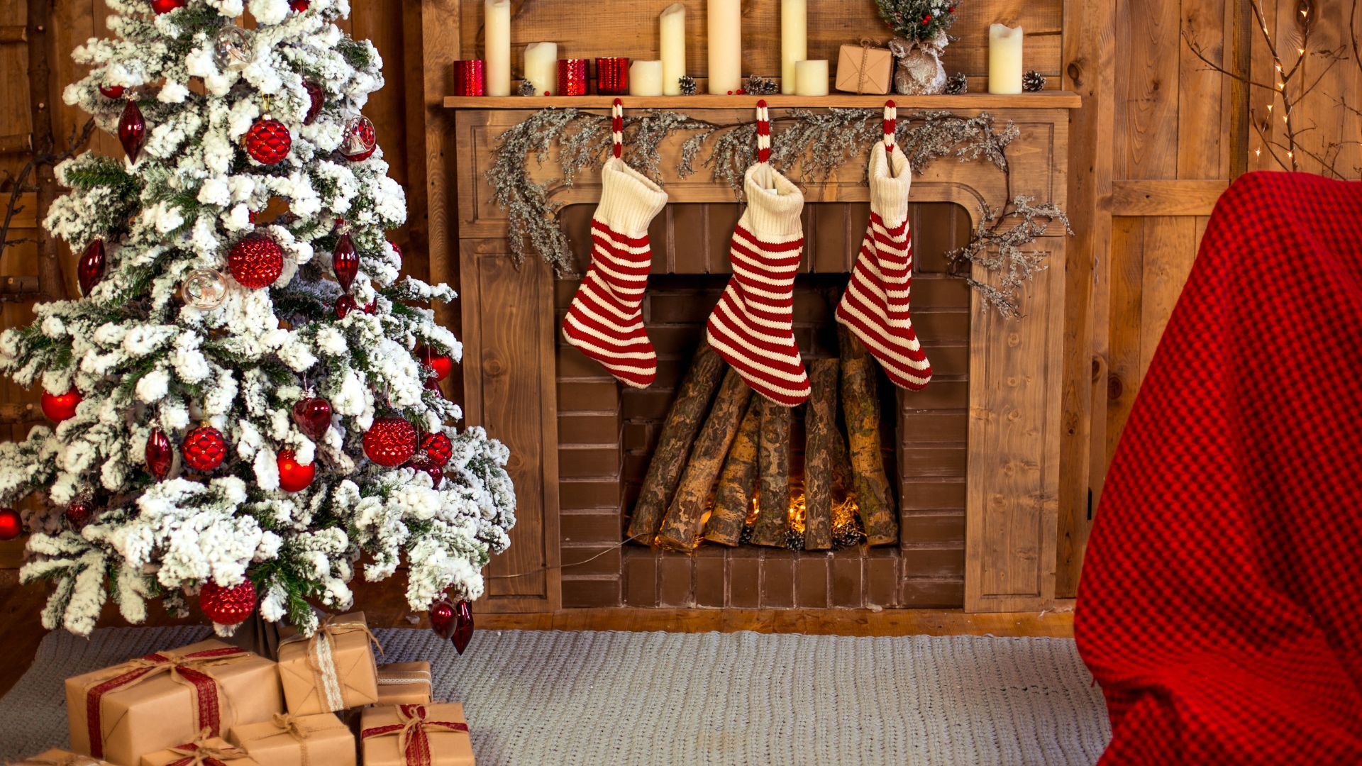 Imagem com uma lareira, decorada com botas natalinas feitas de lã nas cores vermelho e branco. Do lado esquedo, uma árvore de Natal decorada e sobre ela, presentes de Natal.