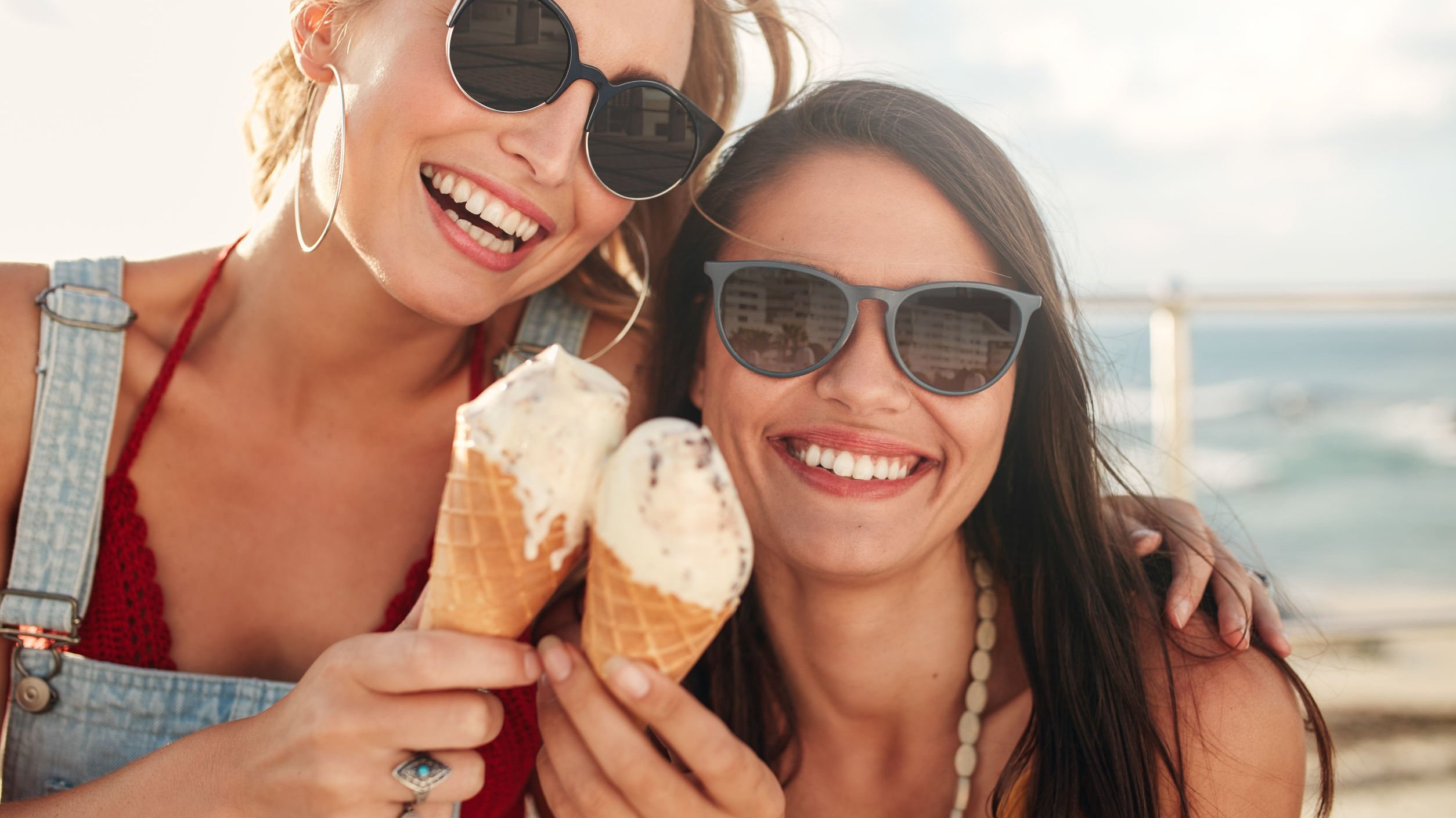 Duas mulheres sorrindo em uma praia segurando sorvete de casquinha