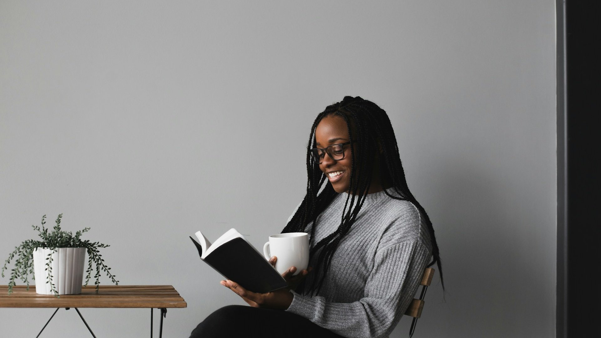 Mulher negra, com cabelos trançados e óculos, lendo um livro. Ela está sentada, sorridente, e segura uma xícara branca na outra mão.