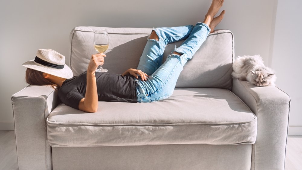 Mulher descansando no sofá com seu gato e, na mão, há uma taça de vinho. Há um chapéu no seu rosto