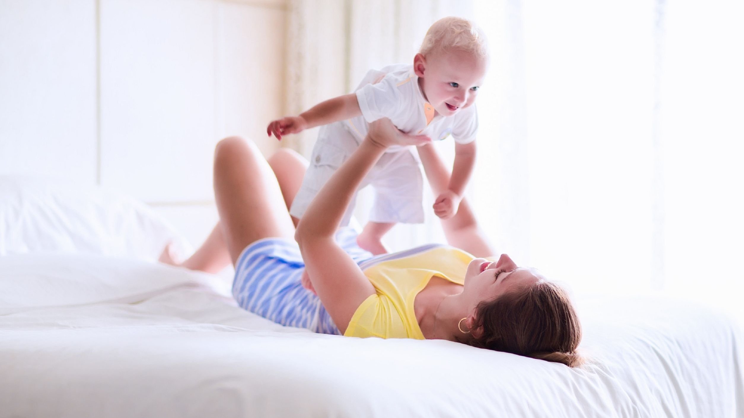 Mulher deitada de barriga para cima em uma cama, segurando um bebê para o alto, que sorri para a mulher.