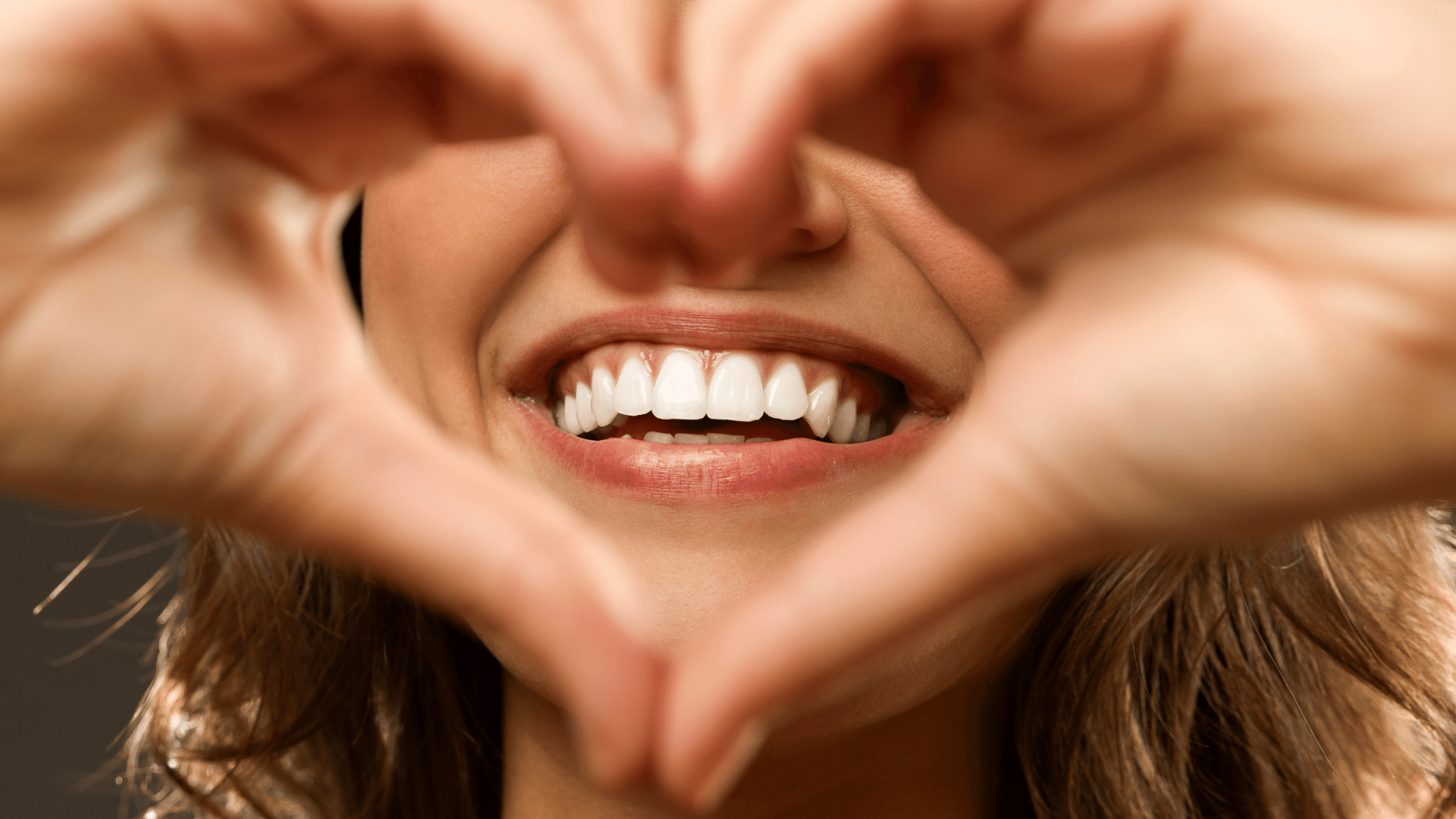 Imagem de uma mulher sorrindo, só mostrando os lábios e com a mão ela fez um coração na frente do sorriso.