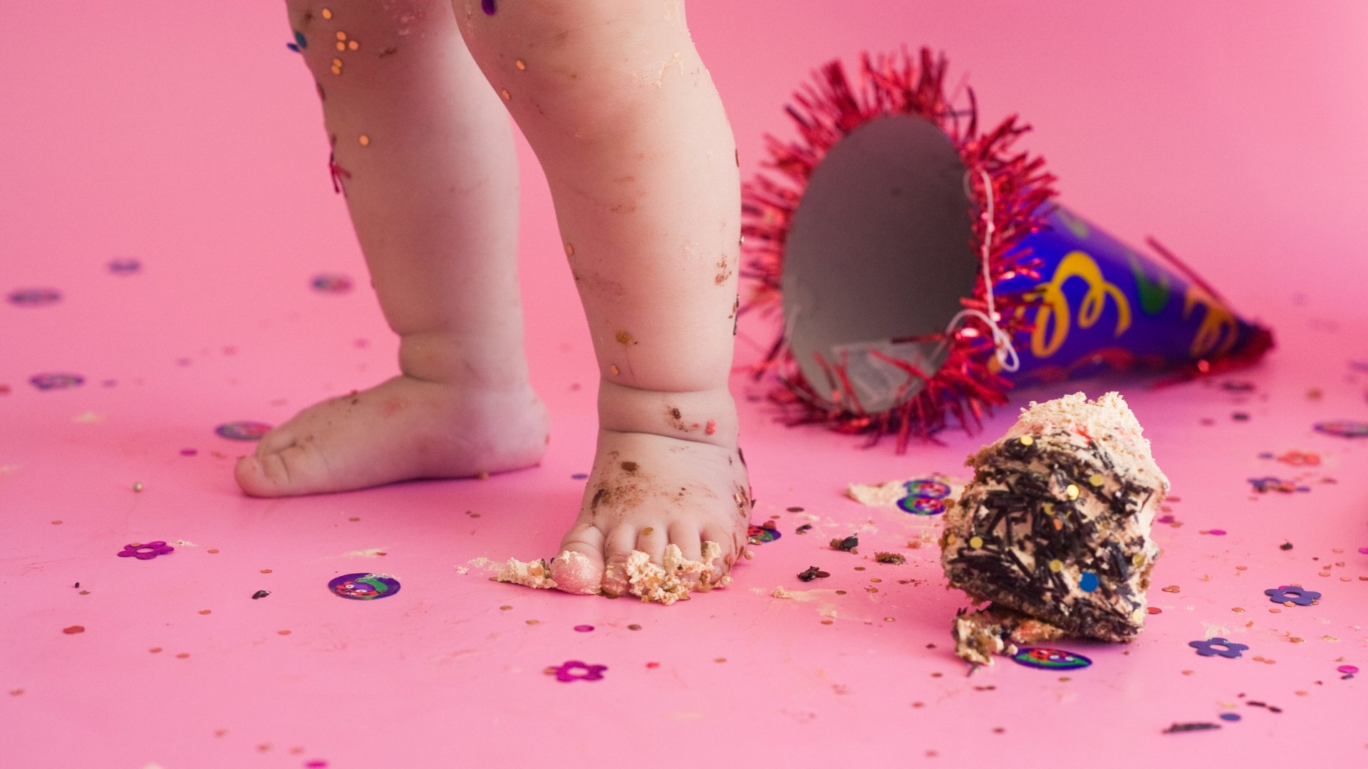 Par de perninhas de bebê sujas de bolo ao lado de um chapéu de aniversário caído no chão de confete com bolo pisado