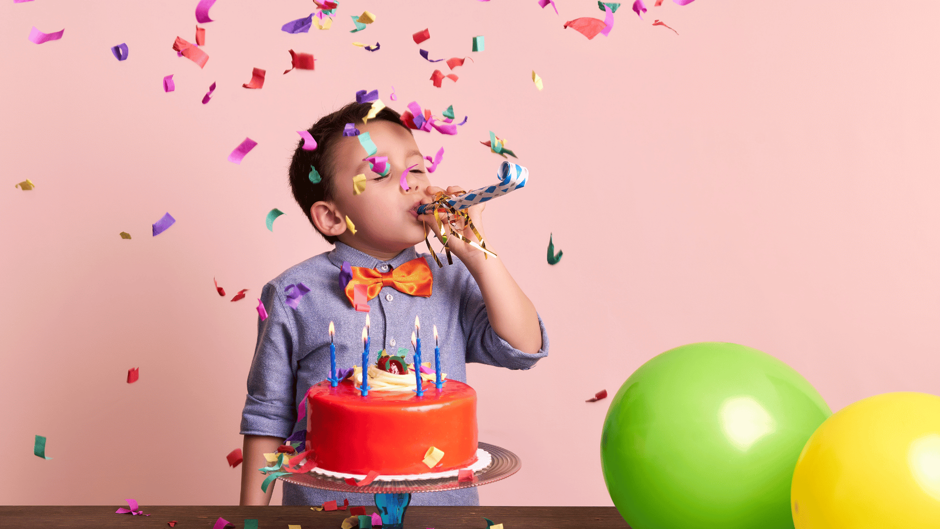 Menino criança assoprando uma língua de sogra próximo de seu bolo de aniversário em um fundo com balões e confetes de festa.