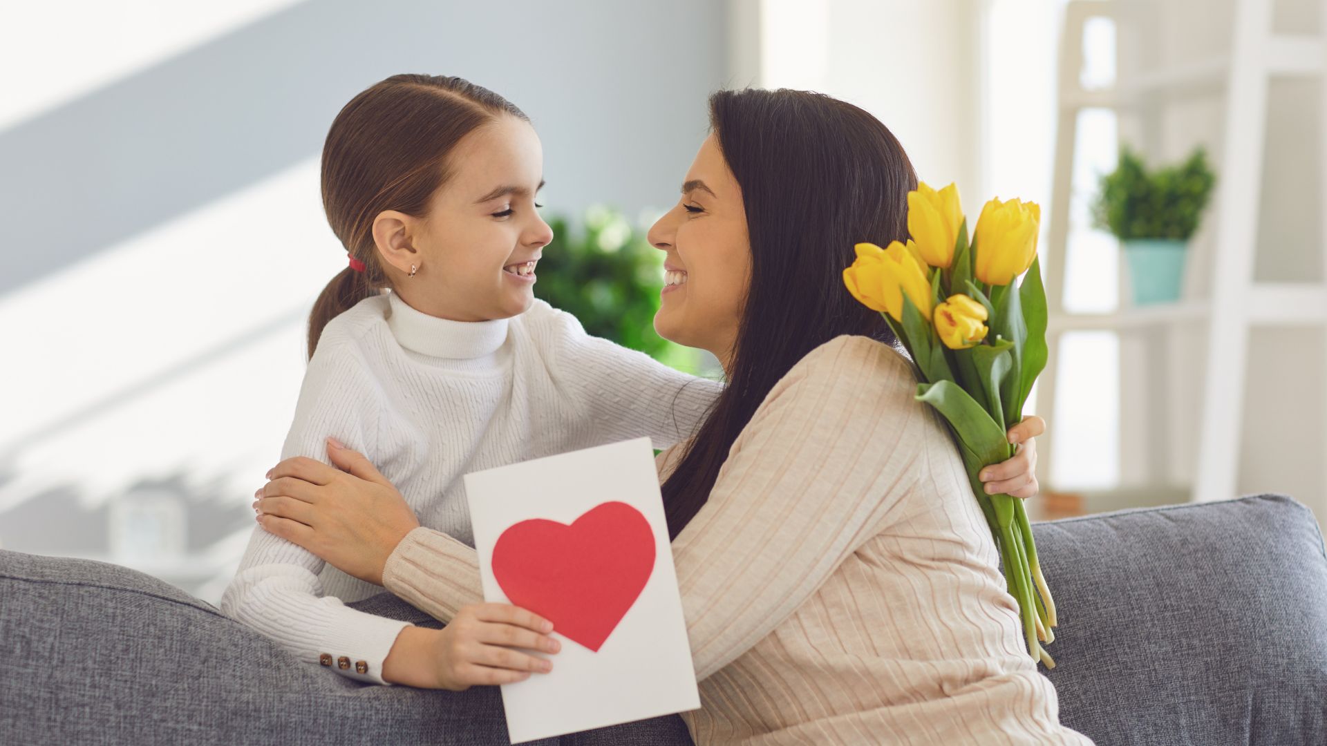 Imagem de mãe e filha se abrançando. A filha entrega um ramalhete de tulipas amarelas e um cartão com um coração desenhado.